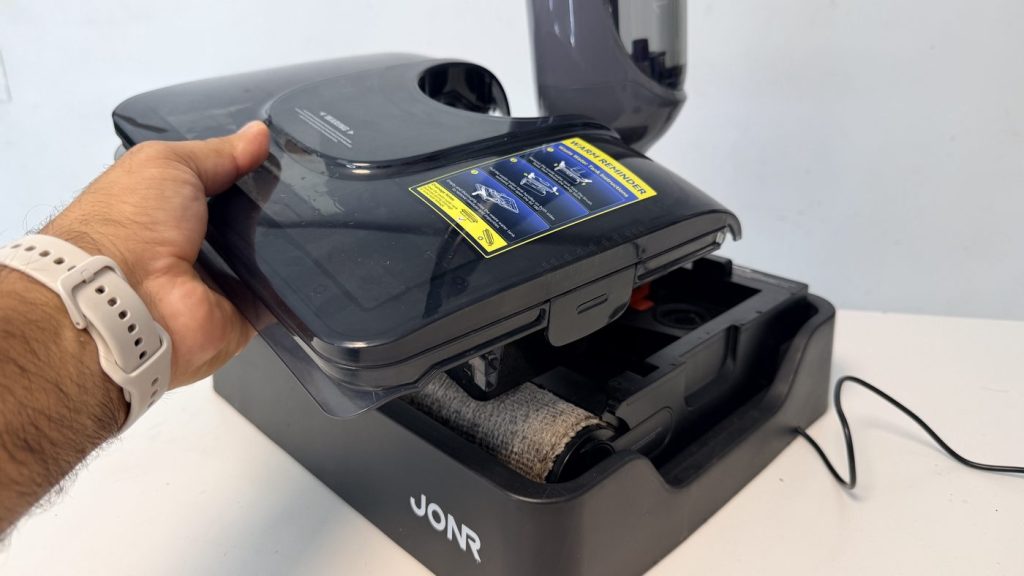 Mano levantando la tapa del depósito de agua sucia del JONR H2 Pro con el rodillo visible debajo y la etiqueta amarilla de aviso